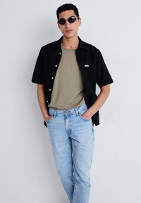 Camisa negra de manga corta con botones sobre una camiseta verde oliva, combinada con jeans de mezclilla azul claro. Modelo con gafas de sol, manos en los bolsillos.