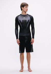 Czarna koszulka z długim rękawem i szarym wzorem, w połączeniu z czarnymi krótkimi spodenkami kąpielowymi z białymi bocznymi paskami. Lekka, dopasowana konstrukcja.