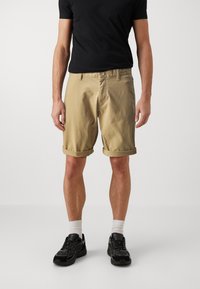 Bermudas de algodón beige con puños doblados, bolsillos frontales y cierre de botón, combinadas con una camiseta negra y zapatillas deportivas negras.