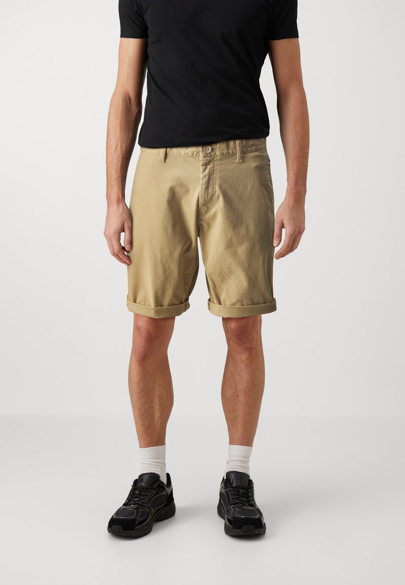 Bermudas de algodón beige con puños doblados, bolsillos frontales y cierre de botón, combinadas con una camiseta negra y zapatillas deportivas negras.