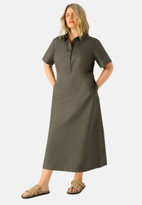Abito verde oliva a maniche corte con colletto, abbottonato davanti e tasche laterali. Indossato con sandali color sabbia, con semplici cinturini e suola piatta.
