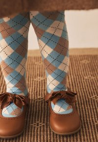 Chaussures en cuir marron avec des accents de nœud papillon, associées à des collants à motif losange bleu, marron et crème. Fond texturé en matériau tissé.