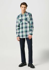 Zielono-niebieska koszula w kratę z dwoma kieszeniami na piersi, długimi rękawami i klasycznym kołnierzykiem, zestawiona z ciemnogranatowymi spodniami i czarnymi butami.