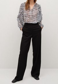 Donna che indossa una blusa a maniche lunghe a fantasia con ruches e pantaloni neri a gamba larga a vita alta, in piedi con le mani nelle tasche.