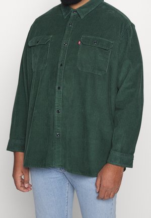 Homme portant une chemise boutonnée en velours côtelé vert foncé avec des poches poitrine et un jean bleu clair, debout devant un fond uni.