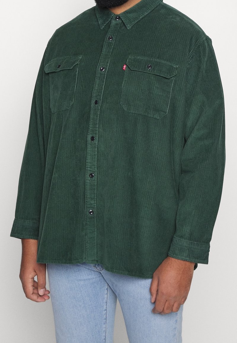 Homme portant une chemise boutonnée en velours côtelé vert foncé avec des poches poitrine et un jean bleu clair, debout devant un fond uni.