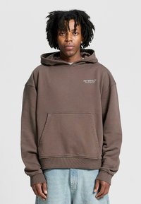 Brown hooded sweatshirt with a front pocket. Features a small logo on the chest. Soft texture, relaxed fit, and ribbed cuffs. Worn with light blue jeans.