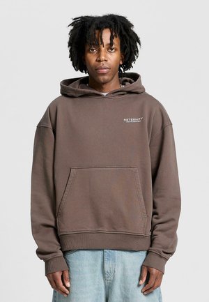 Brown hooded sweatshirt with a front pocket. Features a small logo on the chest. Soft texture, relaxed fit, and ribbed cuffs. Worn with light blue jeans.