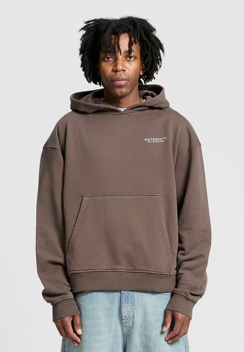 Brown hooded sweatshirt with a front pocket. Features a small logo on the chest. Soft texture, relaxed fit, and ribbed cuffs. Worn with light blue jeans.