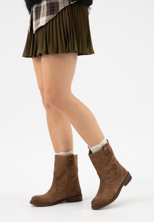 Jambes portant une jupe plissée vert olive, des chaussettes blanches et des bottes en suède marron mi-mollet sur un fond blanc.