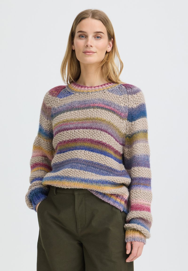Femme aux cheveux blonds mi-longs portant un pull en maille rayé multicolore et un pantalon vert foncé, debout les mains dans les poches.
