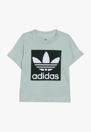 T-shirt vert clair à manches courtes avec un grand carré noir présentant le logo trefoil blanc d'Adidas et le nom de la marque sur le devant.
