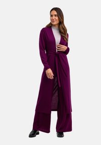 Cardigan long et ouvert en tissu violet profond, doté d'une taille à nouer, associé à un haut gris et un pantalon large. Texture lisse et coupe décontractée.