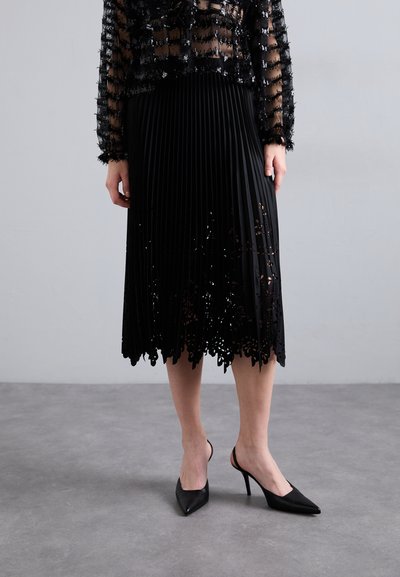 Jupe plissée noire avec une dentelle en bas et des découpes florales, assortie à des escarpins noirs à talons ouverts. Haut noir transparent avec un détail texturé.