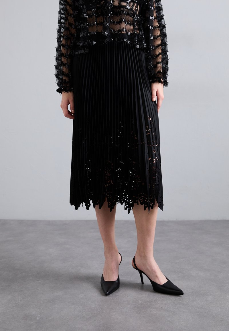 Fekete pliszírozott szoknya csipke szegéllyel és virágos kivágásokkal, fekete, hegyes sarokkal ellátott slingback cipővel párosítva. Áttetsző fekete felső textúrával.