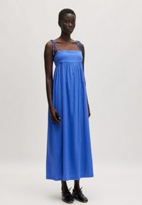 Blaues ärmelloses Maxikleid mit Bindegurten, strukturierter Stoff und gerafftem Bund. Das Model trägt schwarze Schuhe und steht vor einem neutralen Hintergrund.
