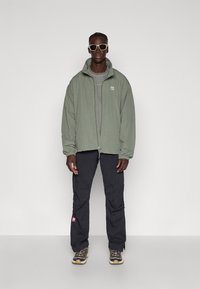 Vaaleanvihreä vetoketjullinen takki, harmaa t-paita, mustat cargo-housut, joissa on punainen logo, ja ruskeat lenkkitossut. Asusteina vaaleat aurinkolasit.