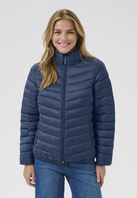 Glunderende vrouw met lang blond haar, gekleed in een navyblauwe gequiltte jas met ritssluiting en blauwe spijkerbroek, tegen een effen grijze achtergrond.