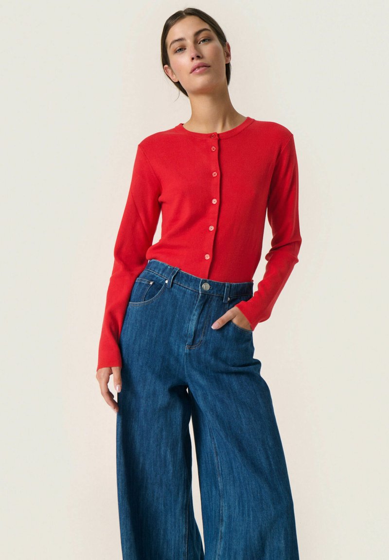 Röd långärmad knapp-up topp i kombination med högmidjade, vida blå denimjeans. Slät textur med en figurnära design och subtila knappar.