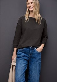 Maglione grigio scuro a coste con maniche ampie, abbinato a jeans in denim blu. La persona tiene una giacca chiara. Sfondo grigio uniforme.