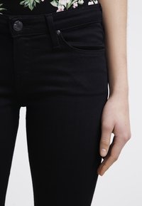 Jeans de mezclilla negra con un ajuste slim, que cuentan con un diseño estándar de cinco bolsillos y un cierre de botón metálico en la cintura. Textura suave.