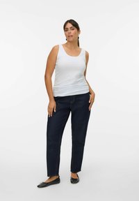 Débardeur blanc à larges bretelles, associé à un jean taille haute bleu foncé. Le mannequin se tient avec une main sur la hanche et porte des ballerines noires.