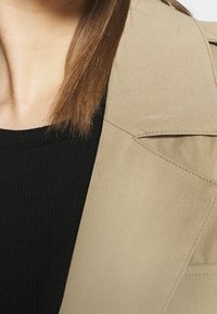 Beżowa marynarka o dopasowanym kroju z wyraźnymi szwami na ramionach, bocznymi kieszeniami i widocznym przeszyciem, zestawiona z czarnym topem w prążki pod spodem.