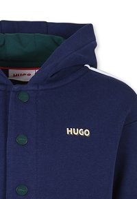 Sweat à capuche bleu marine en tissu doux, avec une doublure intérieure verte, des accents décoratifs blancs et des boutons verts. Comprend le logo "HUGO".