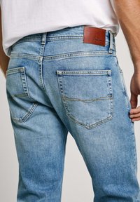 Kiire pilt inimesest, kes kannab heledat sinist denimist teksapüksteid, millel on tagataskud ja pruuni nahkse Pepe Jeans logo silt.