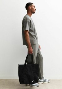 Svart tygväska med två stabila handtag, prydd med en liten vit logotyp. Modell klädd i en grå t-shirt och matchande byxor står bredvid en vägg.