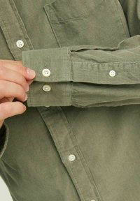 Camicia in corduroy verde con bottoni bianchi e texture rigata, che mostra un polsino abbottonato e una tasca sul petto nella parte anteriore.