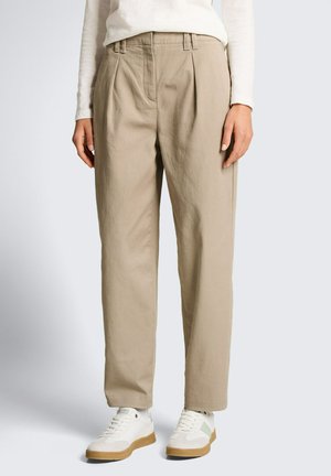 Femme portant un pantalon plissé beige taille haute et des baskets blanches, debout devant un fond clair uni.