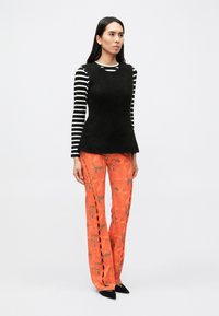 Juodos spalvos rankovių palaidinė dėvima virš juodai baltomis juostelėmis puošto ilgarankovio marškinio, derinama su oranžinės spalvos raštuotomis platėjančiomis kelnėmis ir juodais aukštakulniais bateliais su aštriais galiukais.