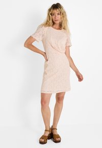 Robe en dentelle rose avec de courtes manches festonnées, silhouette ajustée et longueur au-dessus du genou. Présente un motif texturé et des poches.