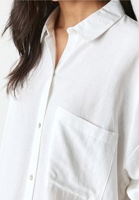 Camicia bianca con bottoni in tessuto leggero, caratterizzata da un colletto a punta e una tasca sul petto. Tessuto liscio con vestibilità comoda.