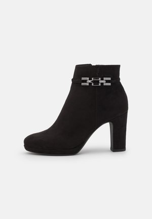 Bottine noire en suède avec un bout arrondi, un haut talon bloc et un accent de chaîne en argent décoratif autour de la cheville. Fermeture éclair sur le côté.