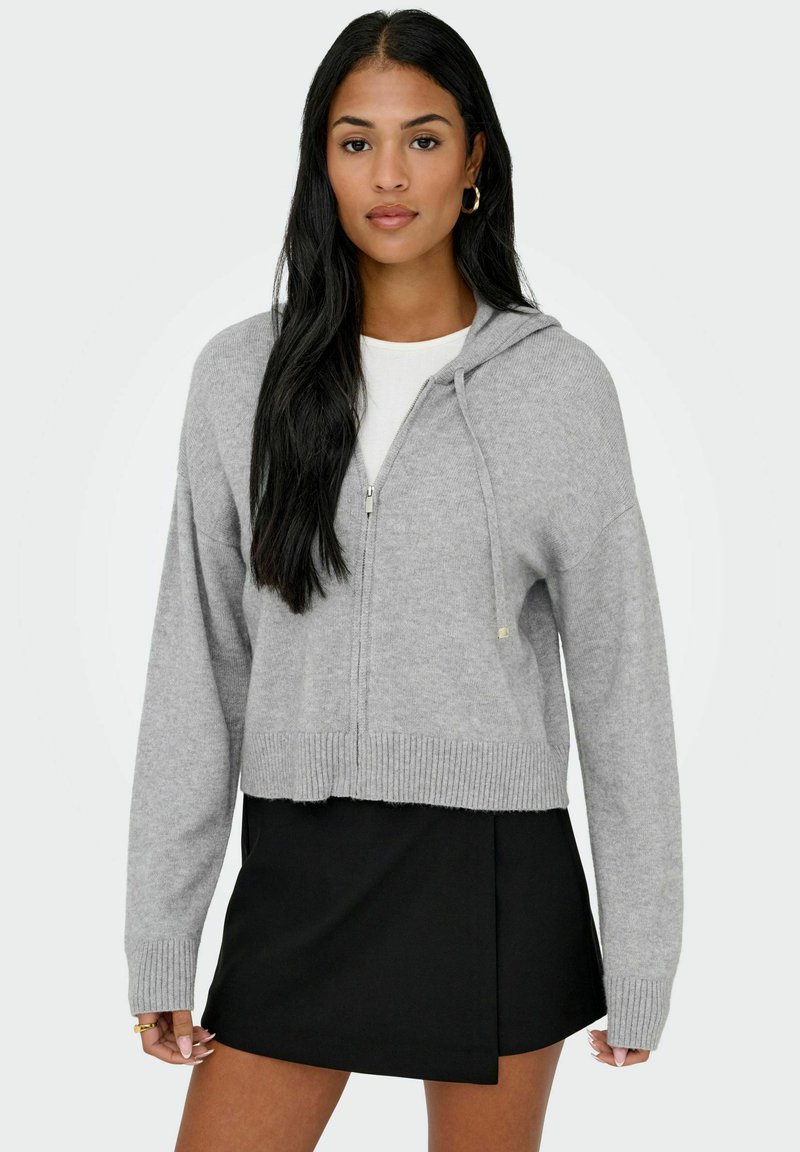 Pelēks īss džemperis, kas izgatavots no mīksta auduma, ar rāvējslēdzēju priekšpusē, ribotiem piedurkņu un apakšmales galiem, kā arī regulējamu kapuci ar šnūrīti. Saskaņots ar melniem svārkiem.