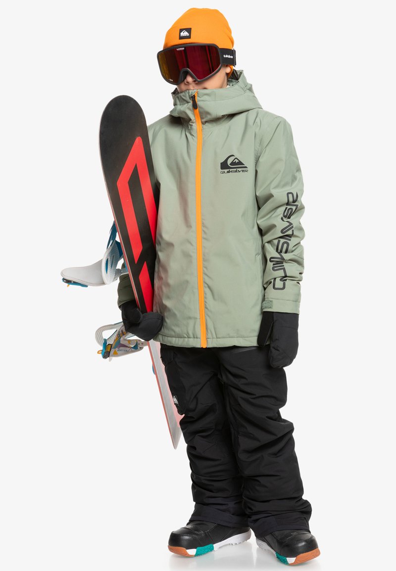 Snowboardista ve světle zelené voděodolné bundě s oranžovým zipem, černých kalhotách, černých rukavicích a oranžovém čepici, držící snowboard.