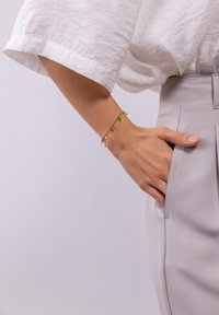 Goldfarbener Armreif mit grünen, orangefarbenen und klaren Anhängern. Trägt eine Hand, die in der Tasche einer hellgrauen Hose ruht. Weiches, strukturiertes weißes Oberteil.