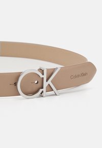 Cintura Calvin Klein in pelle marrone con fibbia circolare CK in argento; texture liscia e design minimalista con marchio impresso.