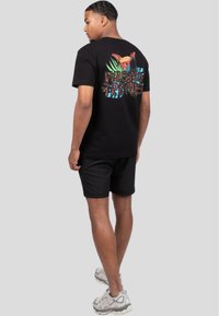 Sort bomulds T-shirt med et farverigt tropisk design, der indeholder en tukan og tekst på ryggen; parret med sorte shorts og lyse sneakers.