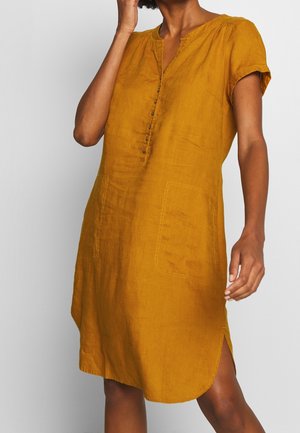 Femme portant une robe en lin jaune moutarde à manches courtes, avec des boutons sur le devant et deux grandes poches frontales.