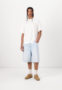 Hvit oversized t-skjorte med et kursiv logo, lys blå denimshorts, brune sneakers og hvite sokker; minimalistisk design, avslappet passform.