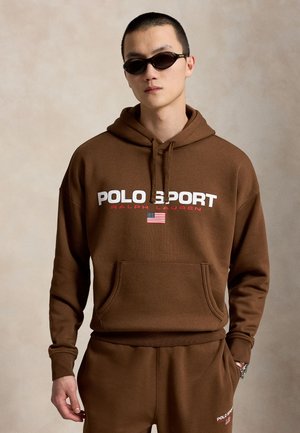 Jeune homme portant un sweat à capuche Polo Sport marron et un pantalon assorti avec un petit drapeau américain, lunettes de soleil foncées et cheveux courts, debout devant un fond uni.