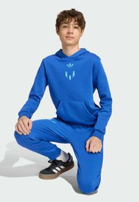 Conjunto de sudadera y pantalones azules hechos de una tela suave. Presenta un bolsillo frontal y un logo en azul más claro. Zapatillas negras con franjas blancas.