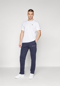White short-sleeve t-shirt with logo on the left chest, paired with navy sweatpants featuring logo on the left thigh. White sneakers worn.