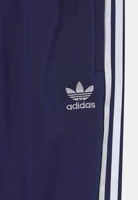 Námořnické sportovní kalhoty mají texturovanou látku, bílé pruhy po stranách a stříbrné vyšívané logo Adidas na horní části levé nohy.