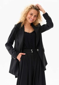 Zwarte blazer met gestructureerde revers, twee voorzakken en decoratieve knoopaccenten. Gemaakt over een zwart top met kantdetails.