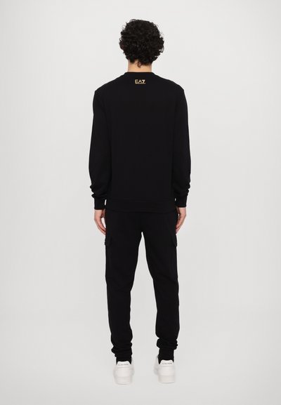Sweatshirt noir à manches longues et bords côtelés, avec un petit logo doré au dos du cou ; associé à un pantalon cargo noir assorti.