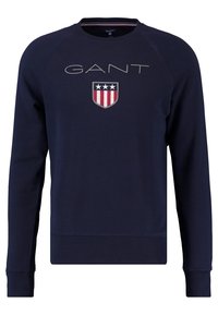 Marineblauwe sweatshirt met raglanmouwen, voorzien van een groot "GANT"-logo en een schild-embleem met rode, witte en blauwe strepen. Katoenmix-stof.